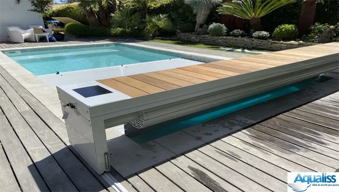 Volet de piscine en forme de banc, de couleur grise, en train de se dérouler sur une piscine à Alès, photographié en journée.