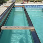 Fixation de la structure porteuse du volet immergé : pose des sabots et des poutres en inox sous les margelles, alignés avec le niveau d'eau.